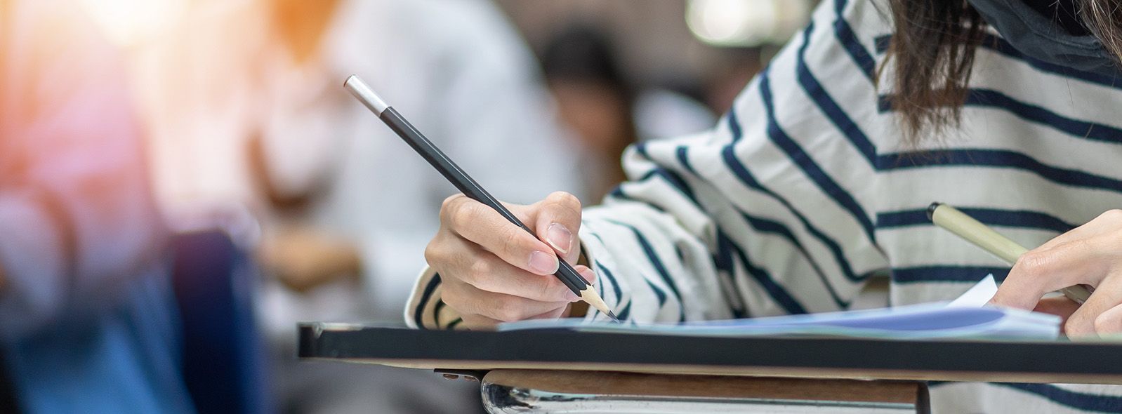 close of of female student writing at a desk in a classroom setting. she is wearing a striped shirt and there are other students in soft focus in the background